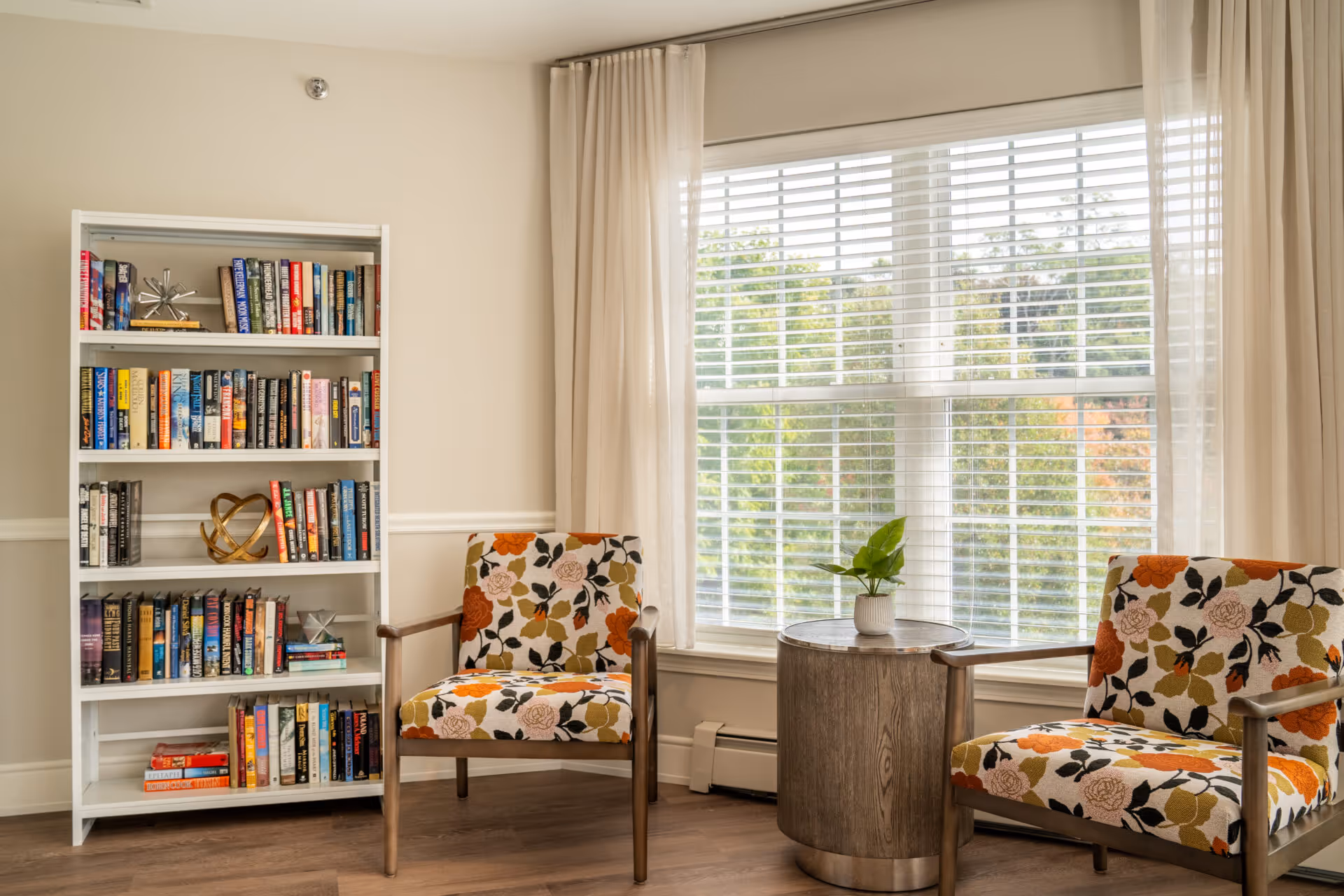 A cozy sitting area with two floral-patterned armchairs facing each other, a round wooden side table with a small potted plant between them, a white bookshelf filled with books and decorative items, and a large window with white blinds and beige curtains letting in natural light.