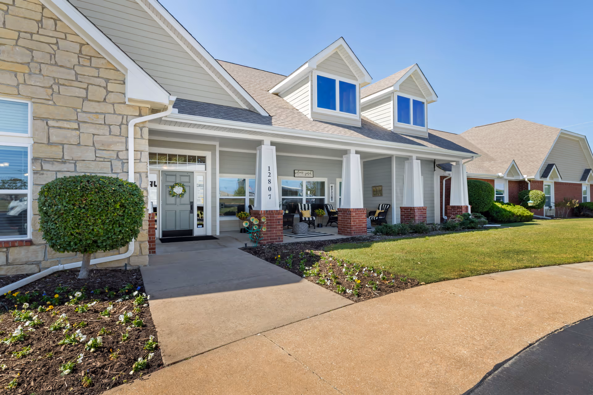 Exterior view of a senior living facility building with a stone and siding facade, a covered porch with seating, manicured bushes, flower beds, and a clear blue sky.