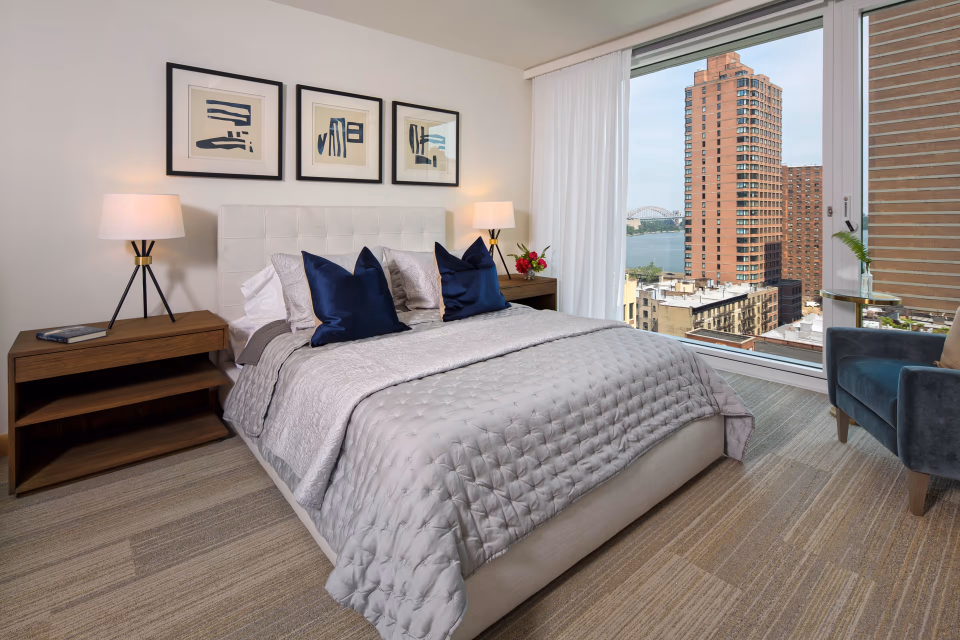 A modern bedroom with a made bed topped with navy pillows, two bedside tables and lamps, framed artwork above the headboard, and a large window showing city and river views.