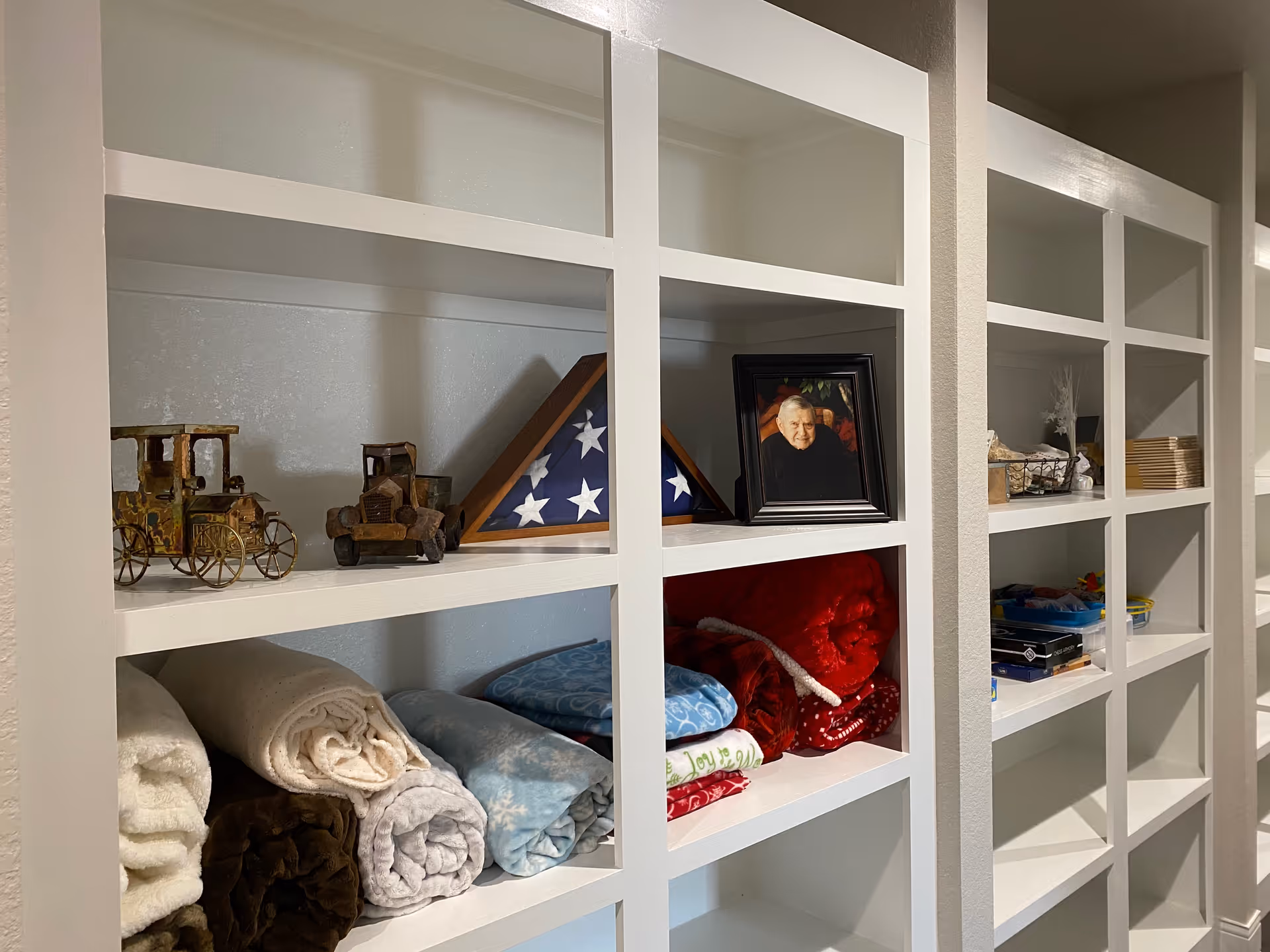 White built-in shelves holding folded blankets, a framed photo of an elderly man, a folded American flag in a triangular display case, small decorative vintage car models, and various other small items.