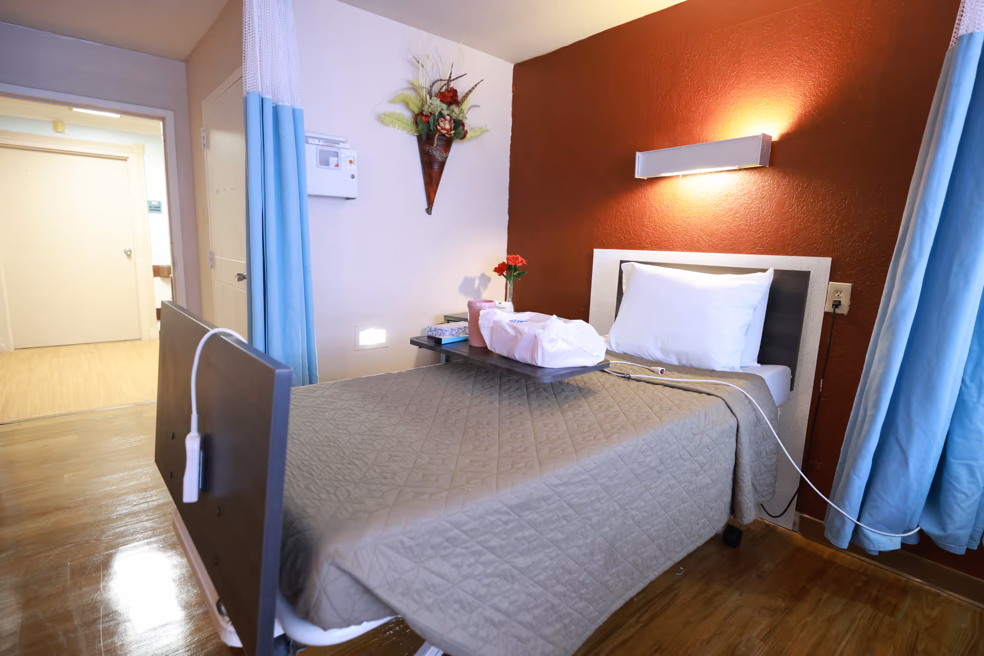 A single hospital-style bed with a gray quilted blanket and white pillow in a room with a red accent wall. A wall-mounted light fixture is above the bed. A small table on the bed holds a wrapped meal and a small vase with red flowers. Blue and white curtains partially frame the bed. The floor is wooden, and there is a doorway leading to a hallway in the background. A decorative wall vase with flowers is mounted on the wall near the bed.