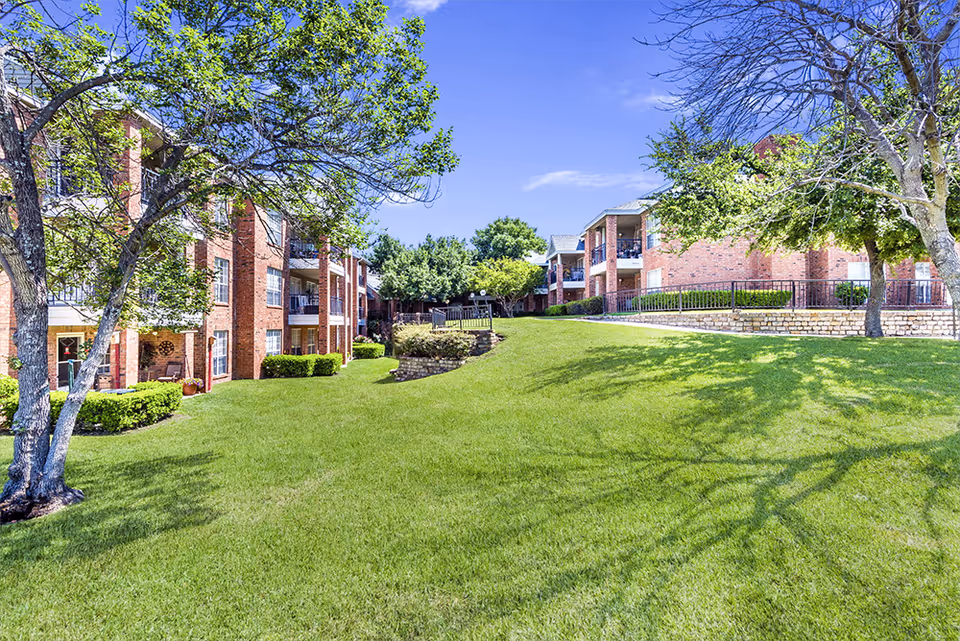 A bright outdoor view of a senior living facility with well-maintained green grass, trees, and brick buildings with balconies under a clear blue sky.
