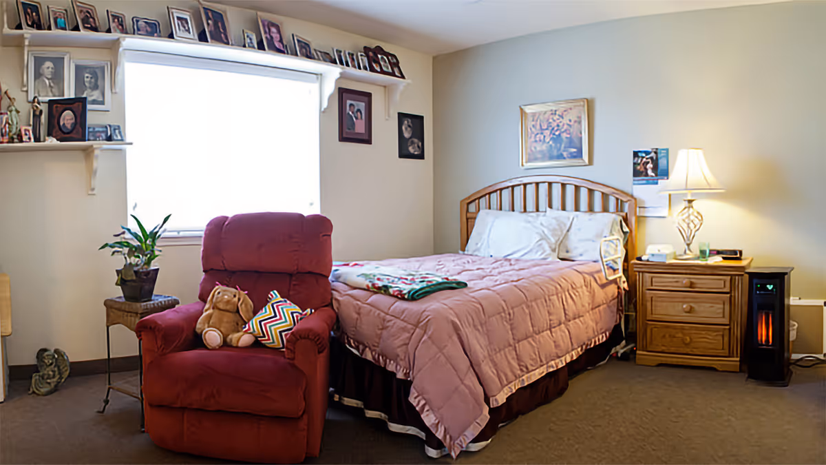 A cozy bedroom with a wooden bed frame and a pink quilted bedspread. Next to the bed is a wooden nightstand with a lamp, a phone, and a small heater on the floor. A red armchair with a stuffed animal and a colorful pillow is placed near a window with white blinds. The walls are decorated with framed photos and artwork, and there is a small table with a plant beside the armchair.