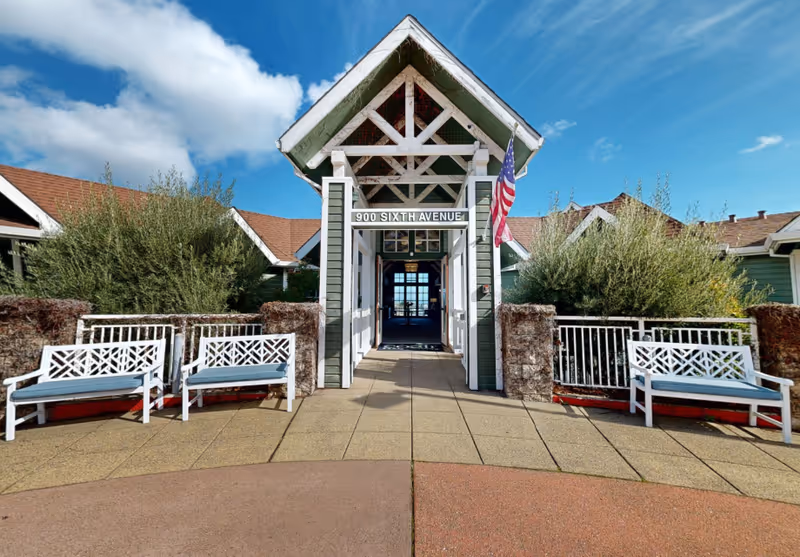 Covered front entrance with a sign reading "900 SIXTH AVENUE", flanked by white benches, greenery, and an American flag under a blue sky.