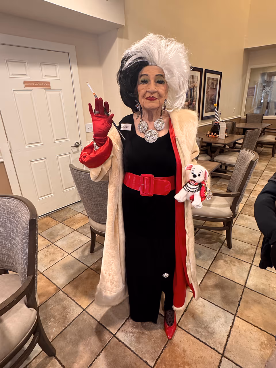 An elderly woman dressed as Cruella de Vil stands in a room with tiled floors and beige walls. She is wearing a black and white wig, a long beige fur coat with a red lining, a black dress with a wide red belt, red gloves, and red shoes. She holds a cigarette holder in one hand and a small stuffed Dalmatian dog in the other. The room has several chairs and tables, framed pictures on the wall, and a door labeled 'Employee Manager Room'.