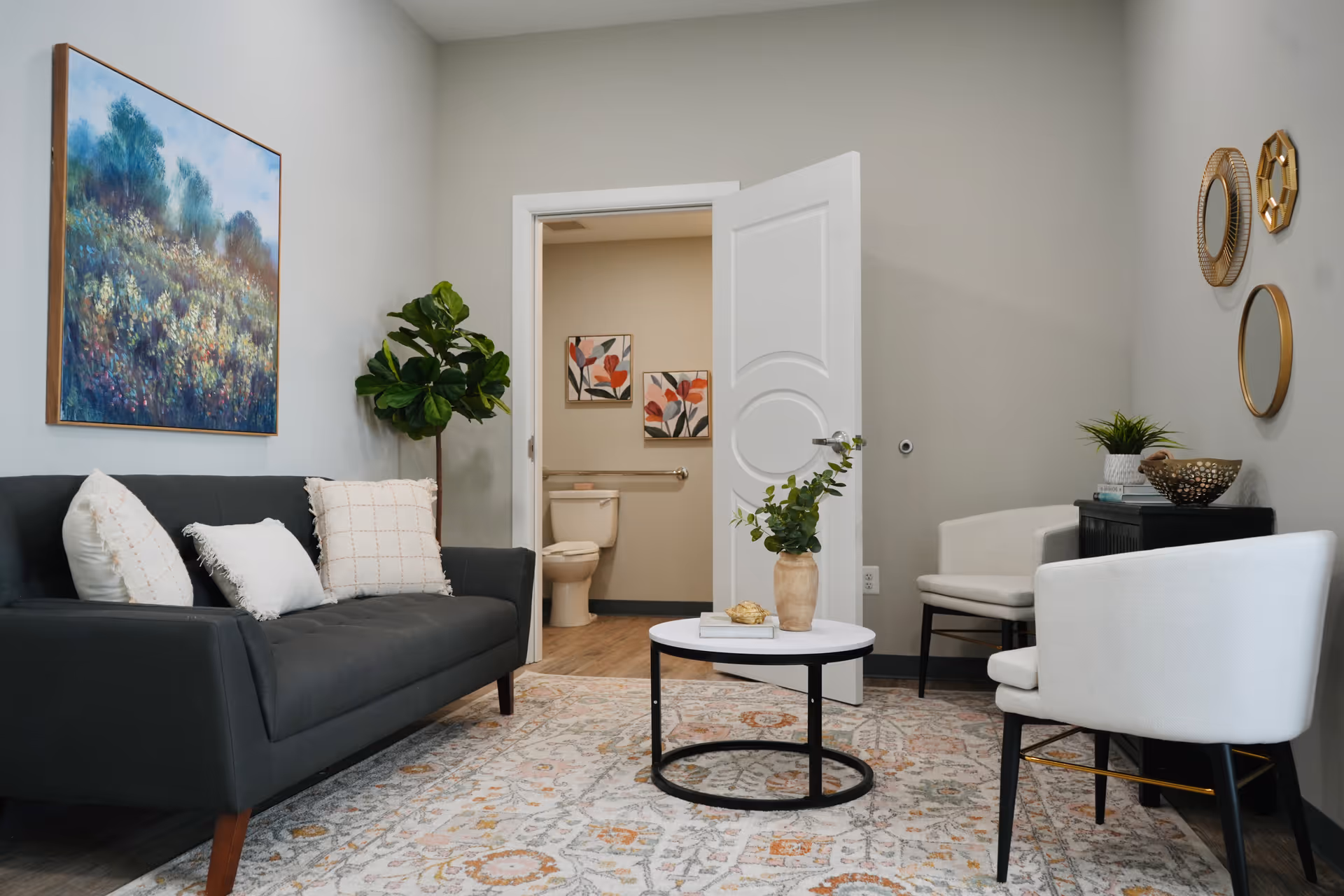 A cozy living room with a dark gray sofa adorned with white pillows, a round white coffee table with a vase of greenery, two white armchairs, and decorative round mirrors on the wall. The room has a patterned rug and a large painting of a floral landscape. An open door reveals a bathroom with a toilet and floral artwork on the wall.
