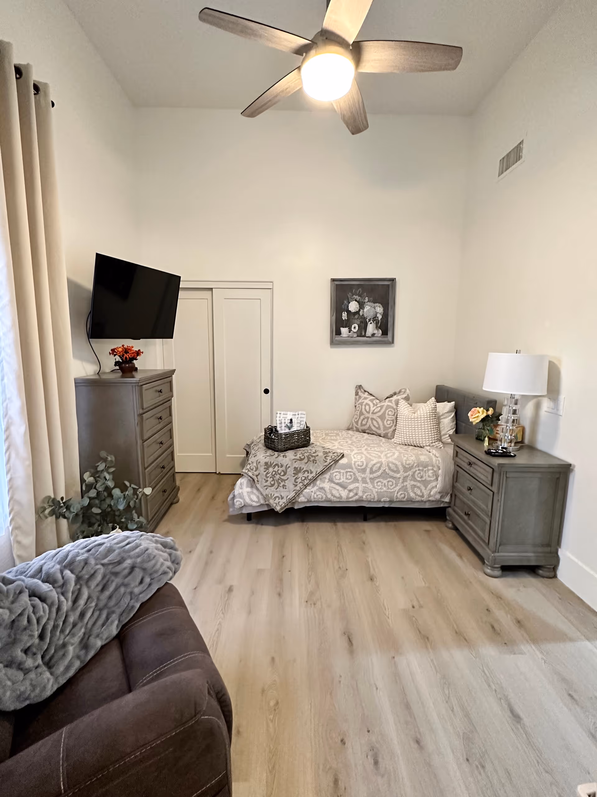 A cozy bedroom with light wood flooring, a single bed with patterned bedding and multiple pillows, a gray nightstand with a lamp and flowers, a gray dresser with a small plant and a wall-mounted TV above it, a brown armchair with a gray throw blanket, beige curtains, a ceiling fan with light, and a framed floral painting on the wall.