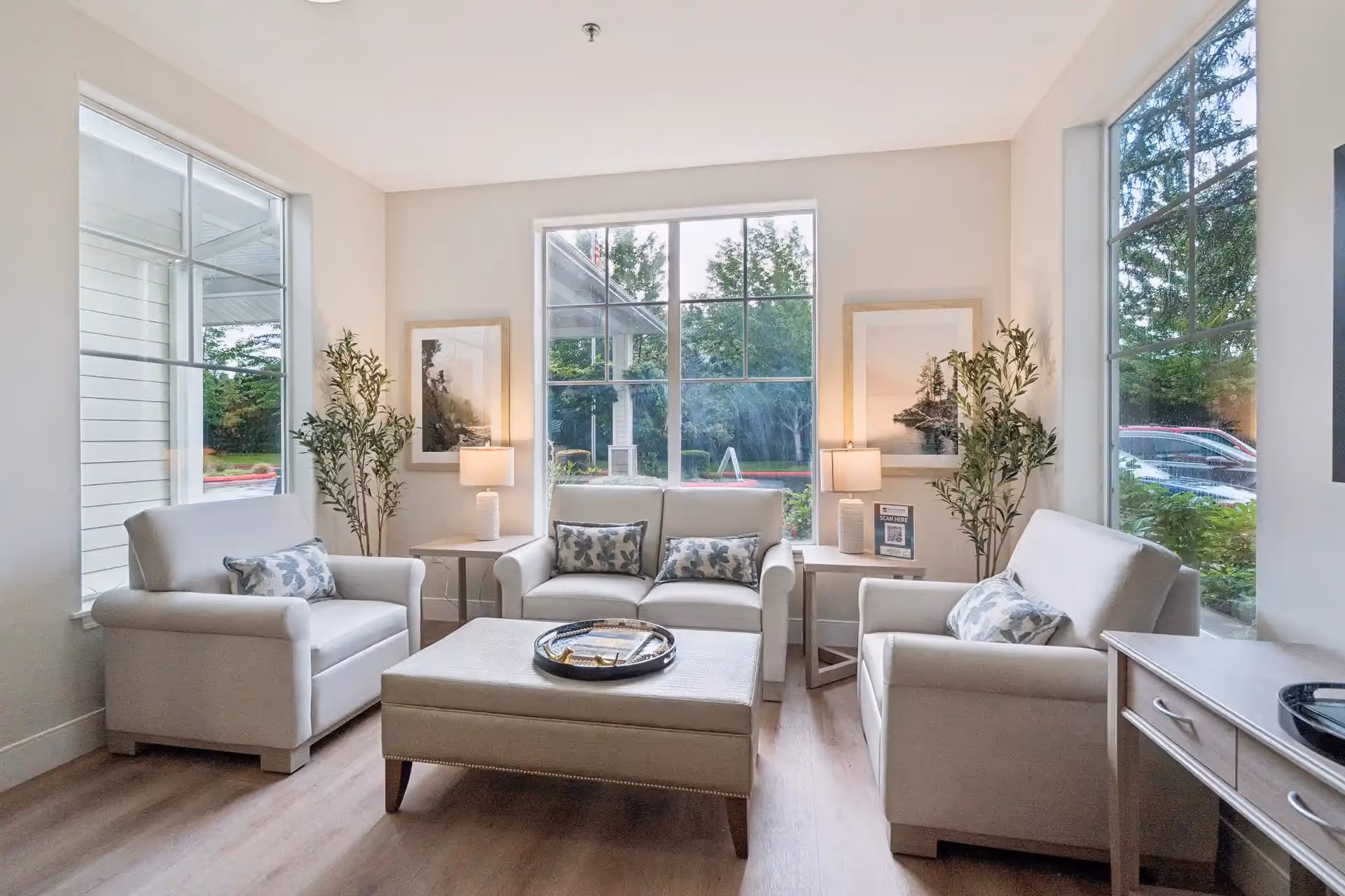 A bright and cozy living room with large windows showing outdoor greenery. The room features two armchairs and a loveseat, all upholstered in light fabric with patterned throw pillows. There are two side tables with lamps and framed artwork on the walls. A large ottoman is centered in the room, and there are decorative plants near the windows.