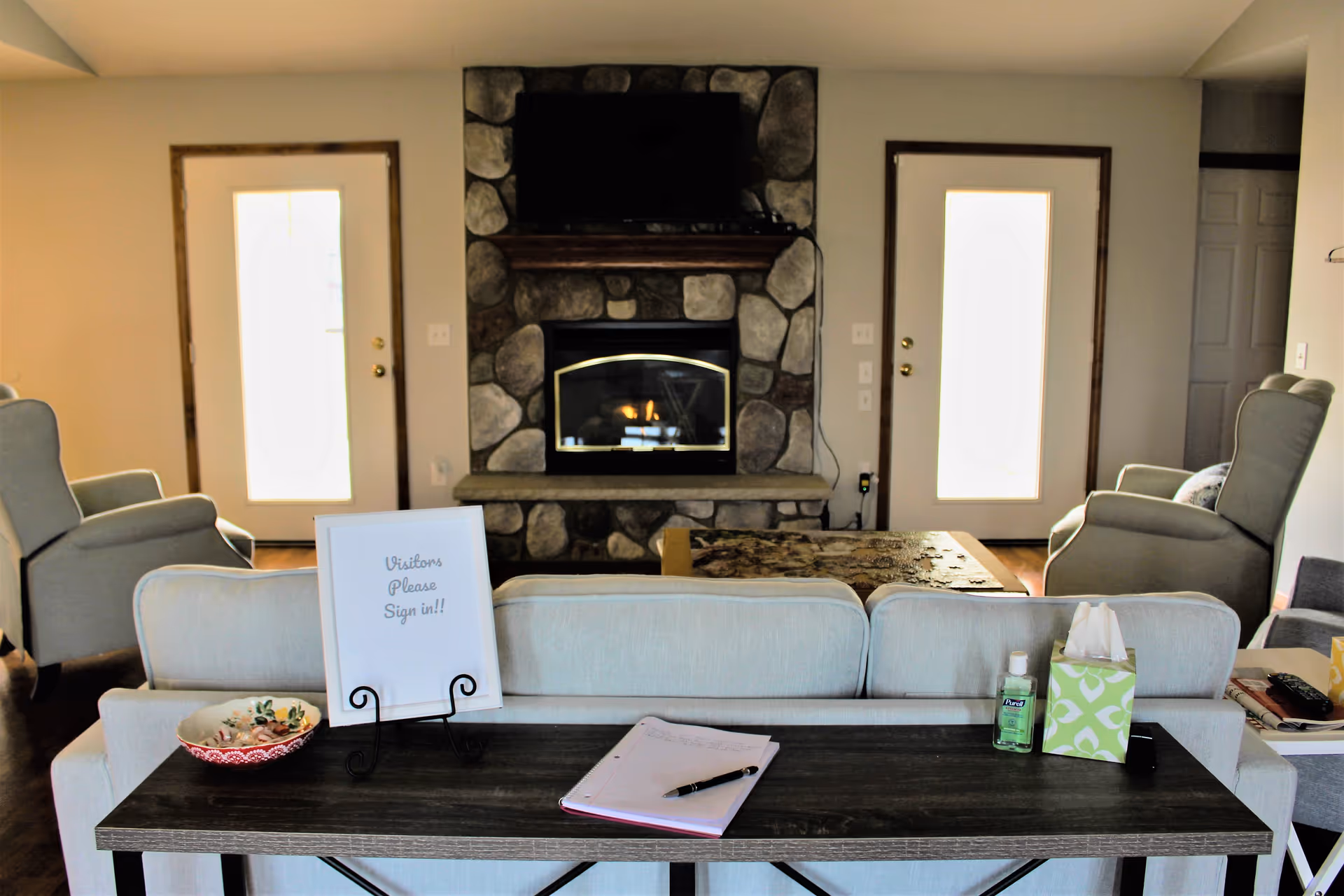 A cozy living room with a stone fireplace and a mounted TV above it. Two armchairs are positioned on either side of the fireplace, and a sofa faces it. Behind the sofa is a table with a sign that reads 'Visitors Please Sign in!!', a notebook with a pen, a bowl, a bottle of hand sanitizer, and a box of tissues. Two doors with frosted glass panels flank the fireplace.