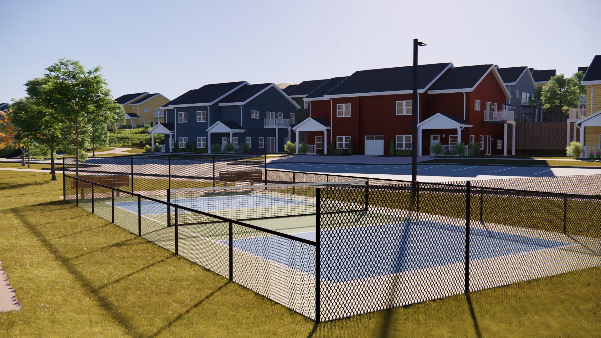 Outdoor fenced pickleball courts with benches around, situated in a grassy area with trees and residential-style buildings in the background under a clear sky.