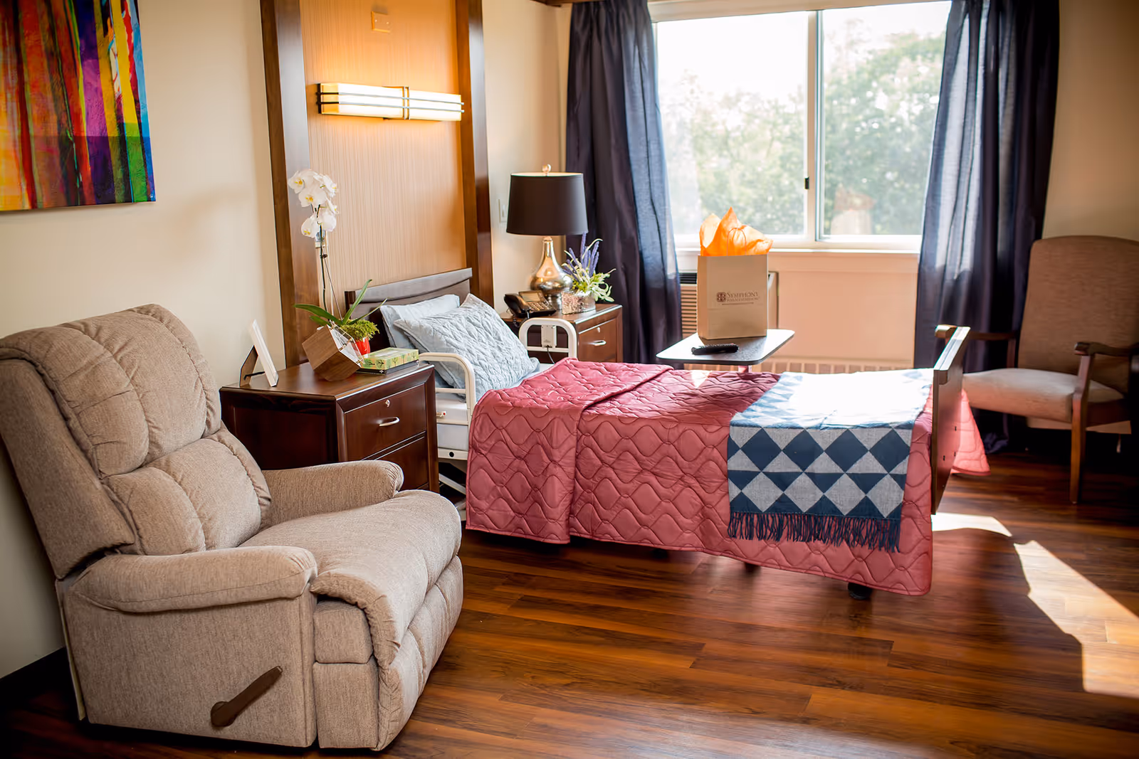 A cozy senior living bedroom with a hospital-style bed covered with a pink quilt and a blue patterned blanket. Next to the bed is a wooden nightstand with a lamp, a phone, and a small plant. There is a beige recliner chair on the left and another armchair near a large window with dark curtains letting in natural light. A colorful abstract painting hangs on the wall.