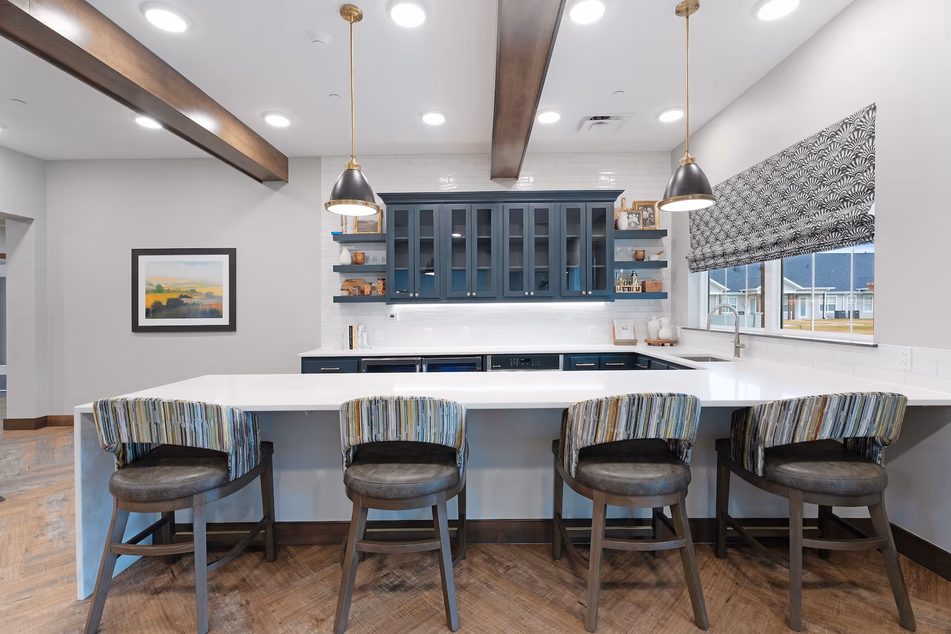 Modern kitchen area with a large white island countertop and four cushioned bar stools with striped backs. The back wall features dark blue cabinets with glass doors, open shelves with decorative items, and a white tiled backsplash. Two pendant lights hang from the ceiling, and a window with a patterned Roman shade is on the right side.