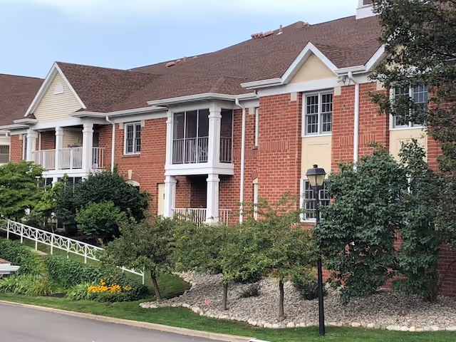 Exterior view of a two-story brick building with white trim and balconies, surrounded by green trees, shrubs, and a well-maintained garden area with rocks and flowers.