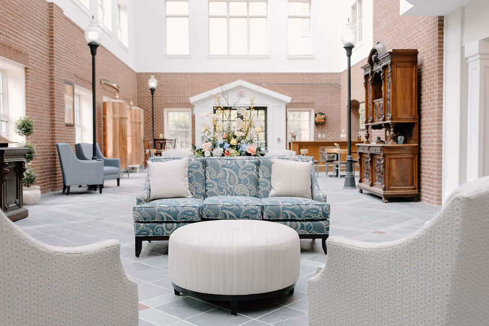 Bright and spacious common area with high ceilings and large windows. The room features a blue patterned sofa with two white pillows, a round white ottoman in front, and two light-colored armchairs in the foreground. There are tall black lamp posts, wooden furniture pieces, and floral arrangements decorating the space. The walls are made of red brick with white trim around the windows and doors.