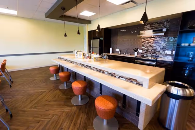 Modern kitchen area with a long beige countertop and five orange cushioned stools. The kitchen features dark cabinetry, a stainless steel refrigerator, stove, and a tiled backsplash with a mix of dark and light tiles. Pendant lights hang above the counter, and there is a trash can to the right side.