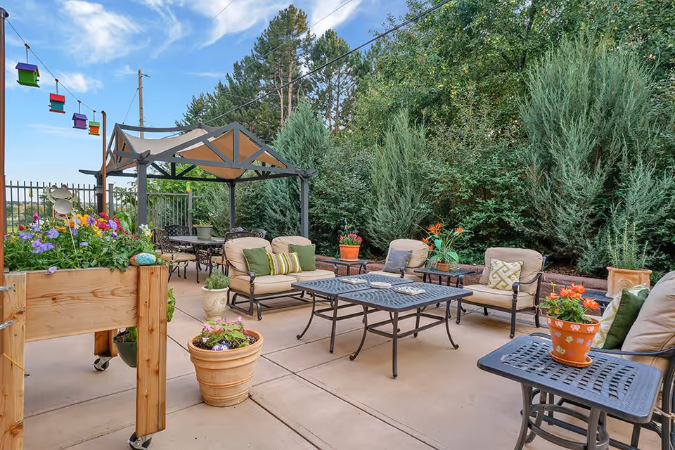 Outdoor patio area with cushioned metal chairs and tables, potted plants, a wooden planter box with colorful flowers, and a canopy structure providing shade. Tall green trees and shrubs form a natural backdrop under a partly cloudy blue sky.