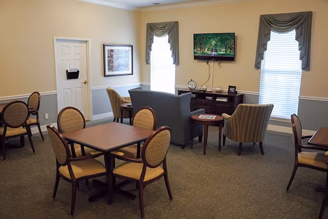 A senior living common room with dining tables and chairs, a seating area of a sofa and armchairs, and a wall-mounted TV between two windows.
