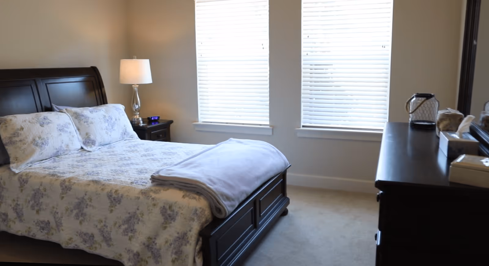 Sunlit bedroom with a dark wooden bed dressed in floral bedding, a nightstand with a lamp, and a dresser under two windows.