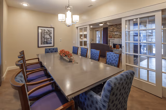 A dining room with a long rectangular wooden table surrounded by blue upholstered chairs. A modern chandelier hangs above the table, and a framed picture is on the beige wall. Sliding glass doors lead to a living room area with a stone fireplace and seating.