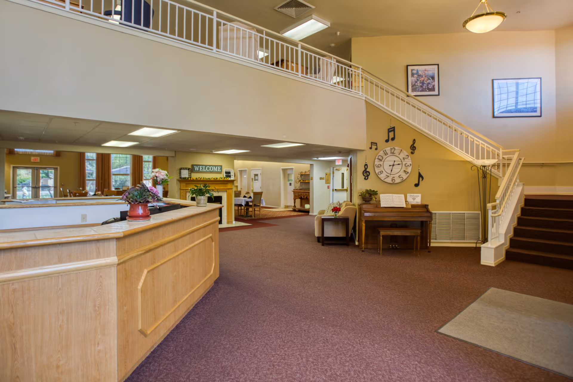 Interior view of a senior living facility lobby area with a wooden reception desk on the left, a staircase with white railings on the right, a piano with musical note decorations and a large clock on the wall, and a seating area with a small table and plant. The space has carpeted floors and warm lighting.