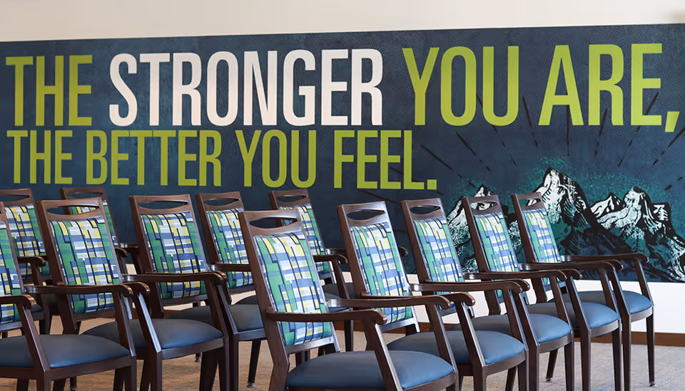 Rows of wooden chairs with patterned cushions in a senior living common area in front of a wall mural that reads "The stronger you are, the better you feel."