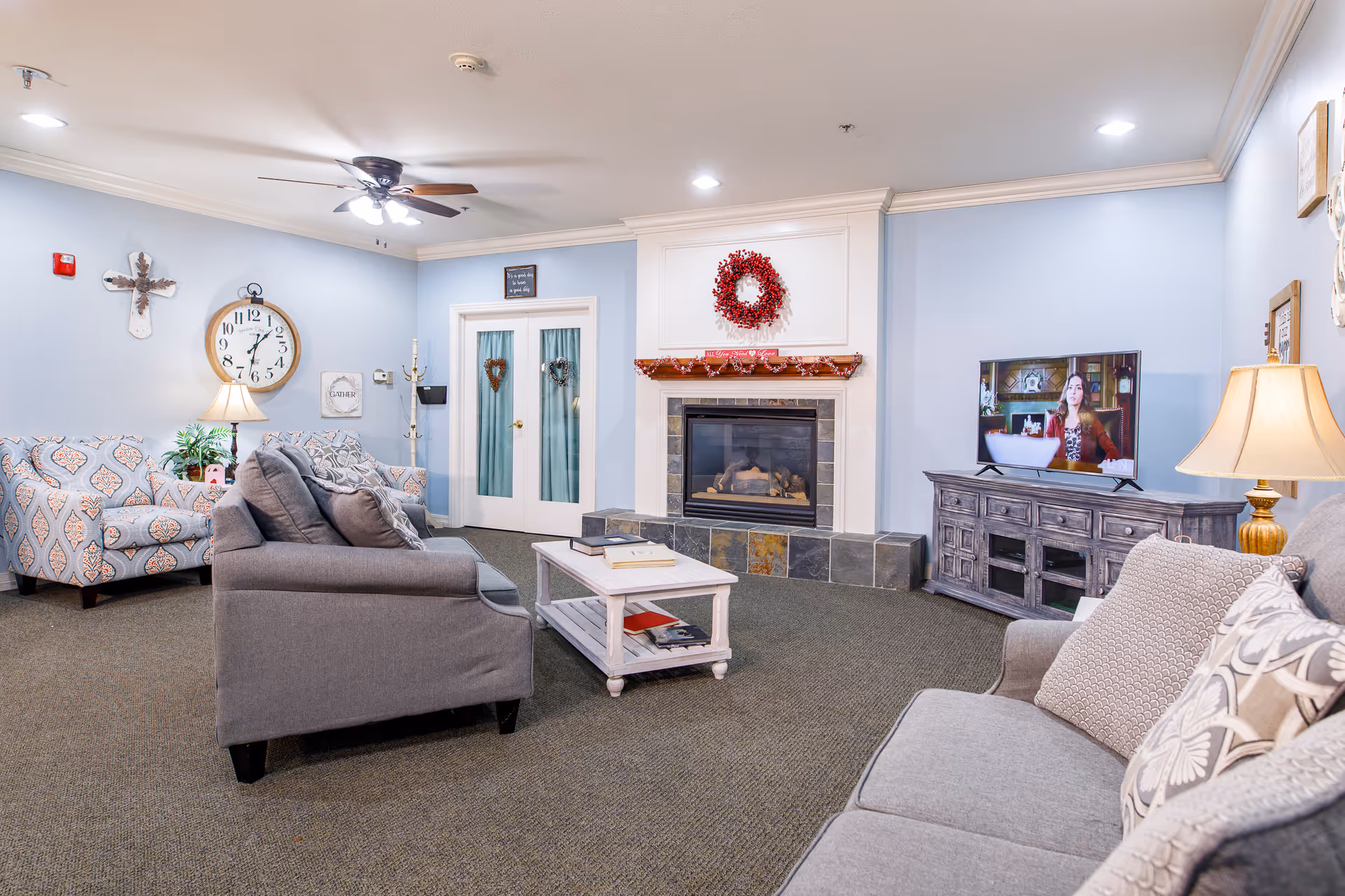 A cozy living room with light blue walls, a ceiling fan with lights, and a fireplace decorated with a red wreath and garland. The room features a gray sofa with patterned pillows, a patterned armchair, a white coffee table with books, a wooden TV stand with a flat-screen TV showing a woman, and various wall decorations including a large clock and a cross.