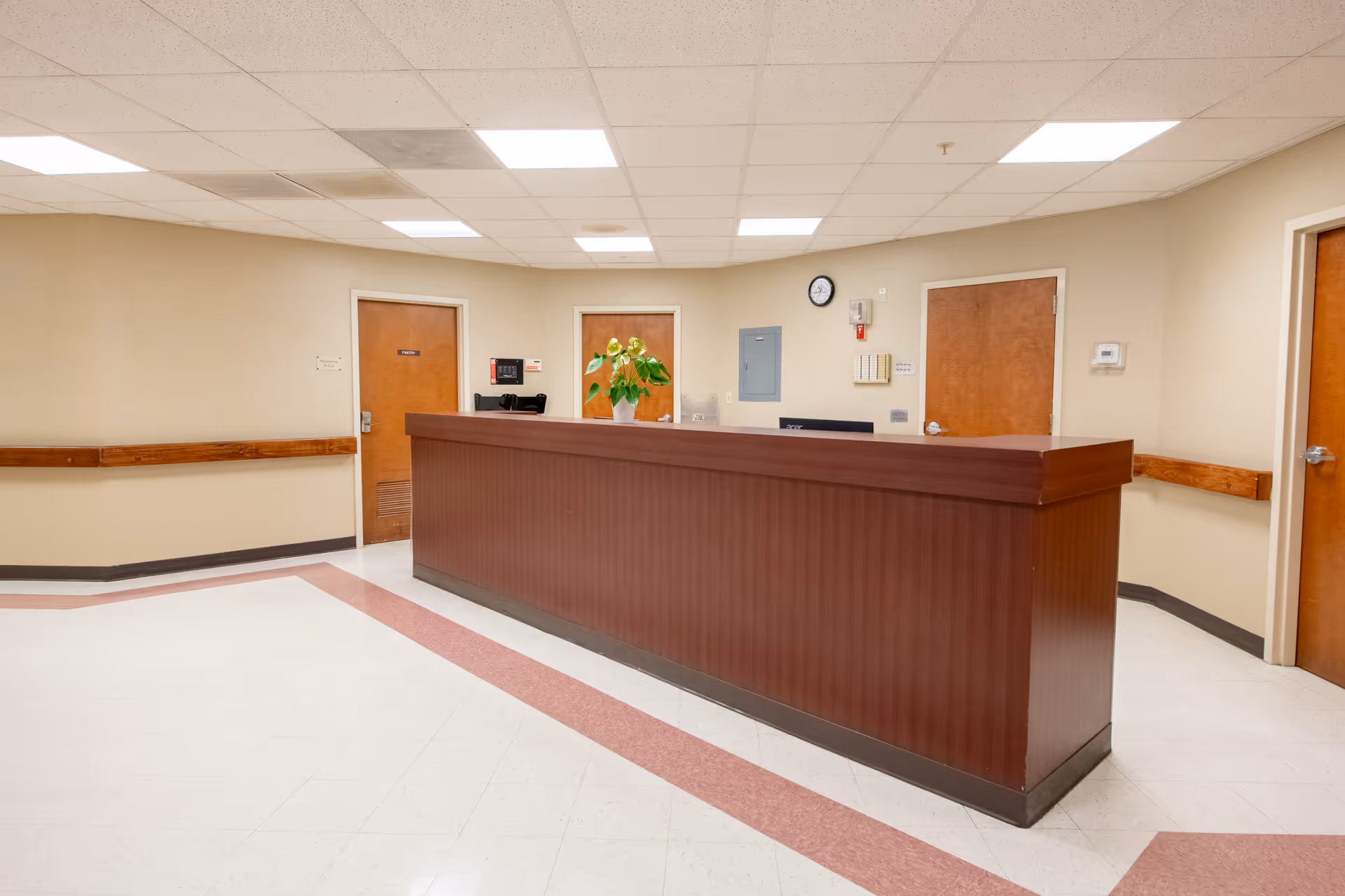 Reception desk area in a healthcare facility with beige walls, a wooden counter, three closed wooden doors, a clock on the wall, and a potted plant on the desk.