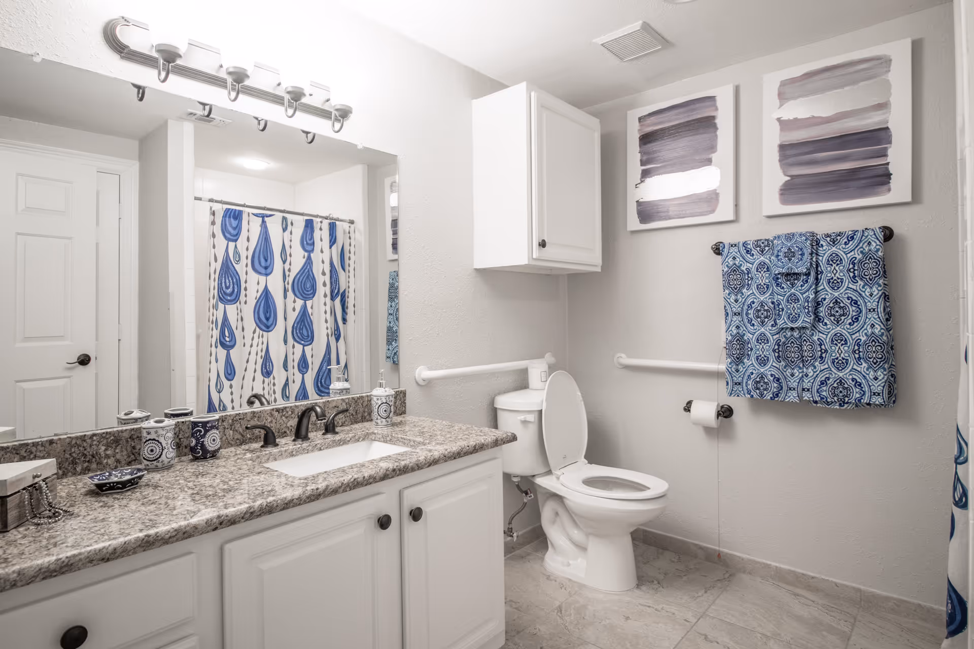 A clean and well-lit bathroom with a granite countertop sink, white cabinets, a toilet with the lid open, a blue and white patterned shower curtain, blue patterned towels hanging on a towel rack, and two abstract paintings on the wall.