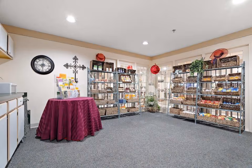 Interior room with carpeted floor featuring metal shelves stocked with various snacks and items in wicker baskets. A round table covered with a burgundy tablecloth holds brochures. Wall decorations include a clock and a decorative cross. The room has beige walls and ceiling lights.