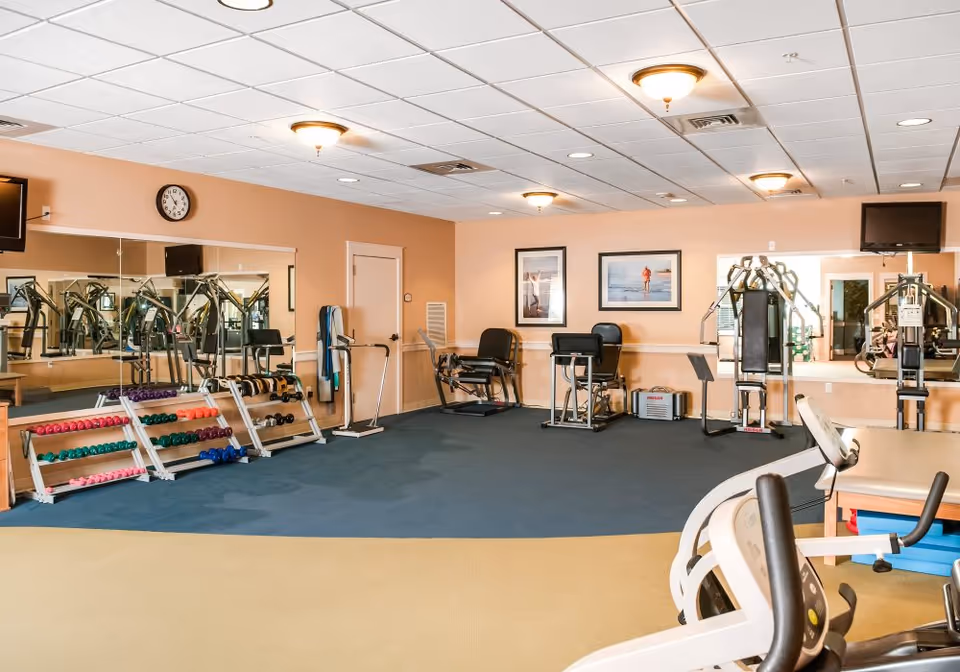 Interior fitness room with weight racks, cardio and resistance machines, mirrors and framed artwork on the walls.