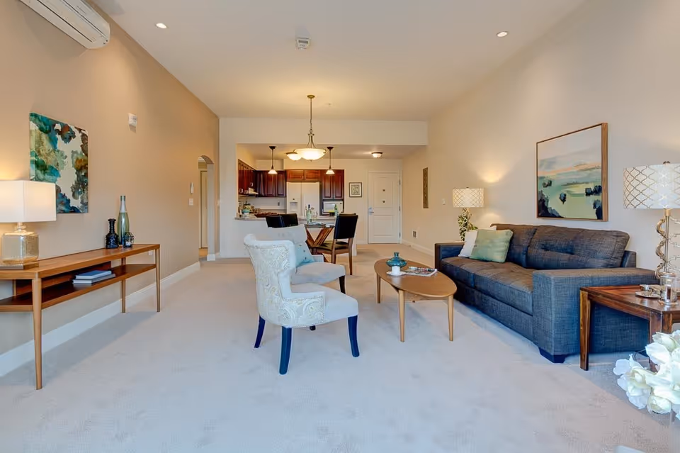 A bright and spacious living room in a retirement community apartment featuring a gray sofa with green and white pillows, two patterned armchairs, a wooden coffee table, and side tables with lamps. The room has beige walls, carpeted floors, and artwork on the walls. In the background, there is a dining area with a table and chairs, and a kitchen with dark wood cabinets and pendant lighting.