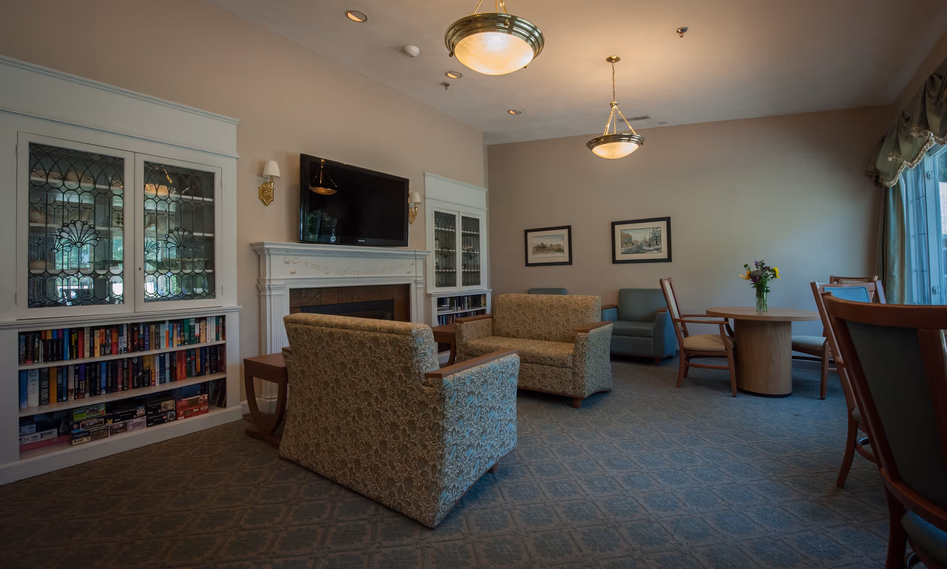 A cozy living room area in a senior living facility with patterned armchairs and a loveseat arranged around a fireplace with a mounted flat-screen TV above it. There are built-in bookshelves filled with books and board games on either side of the fireplace. A round wooden table with chairs and a vase of flowers is near a window with draped curtains. The room is softly lit by ceiling lights and wall sconces.