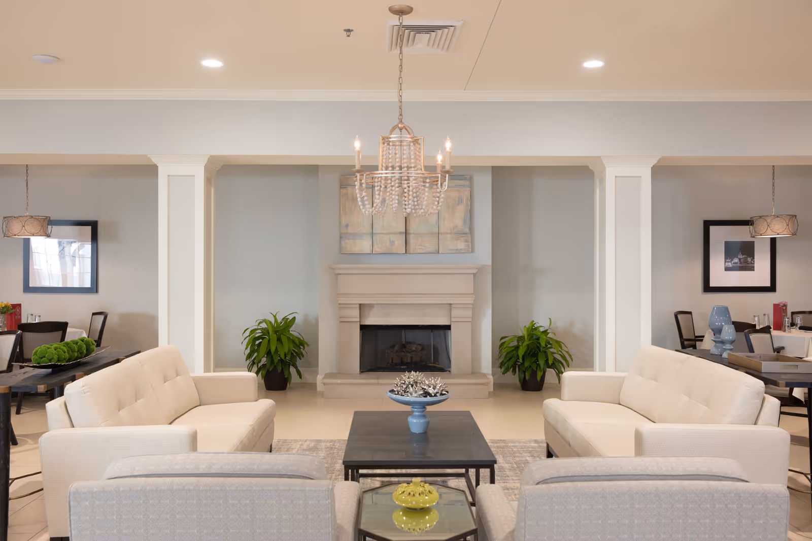 Bright furnished common living room with cream sofas arranged around a central coffee table and a fireplace beneath a chandelier.