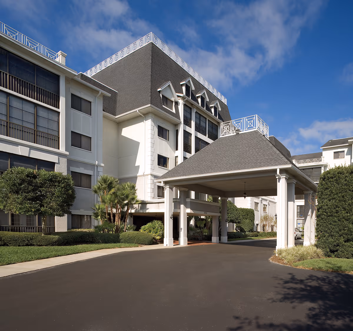 Exterior view of a multi-story senior living facility with a covered entrance driveway, manicured bushes, and clear blue sky.