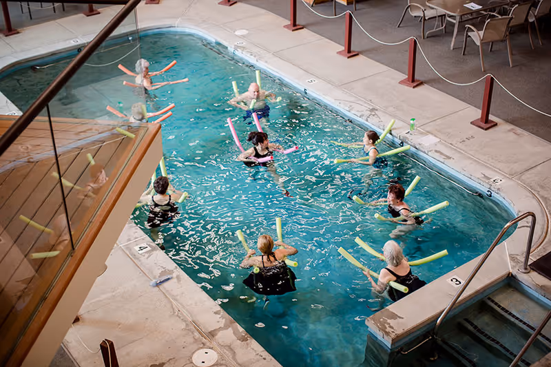 A group of elderly people participating in a water exercise class in an indoor swimming pool, each using colorful pool noodles. The pool area has a tiled deck with chairs and tables nearby.