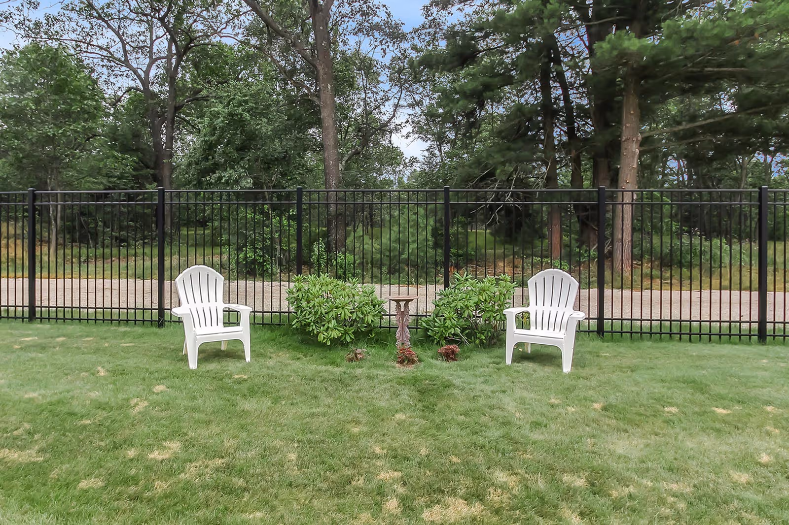 Two white plastic chairs on a grassy lawn facing a black metal fence with green bushes and trees in the background under a cloudy sky.