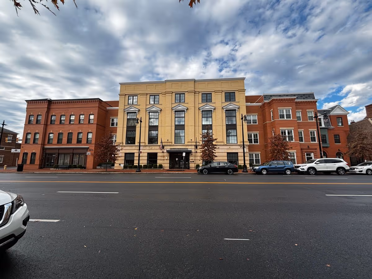 Street-facing facade of a multi-story brick and yellow building with parked cars in front under a cloudy sky.