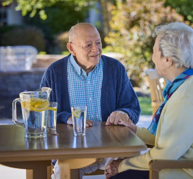 An elderly man and woman sitting at a wooden table outdoors, holding hands and smiling at each other. On the table, there is a pitcher and glasses of water with lemon slices. The background shows greenery and a sunny day.