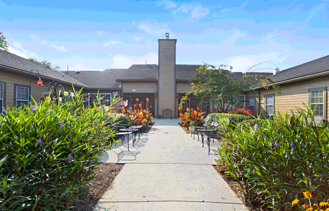 Outdoor courtyard area at American House Shallowford featuring a paved walkway lined with green plants and colorful flowers. There are black metal chairs and tables on either side of the path, with a building in the background under a blue sky with some clouds.