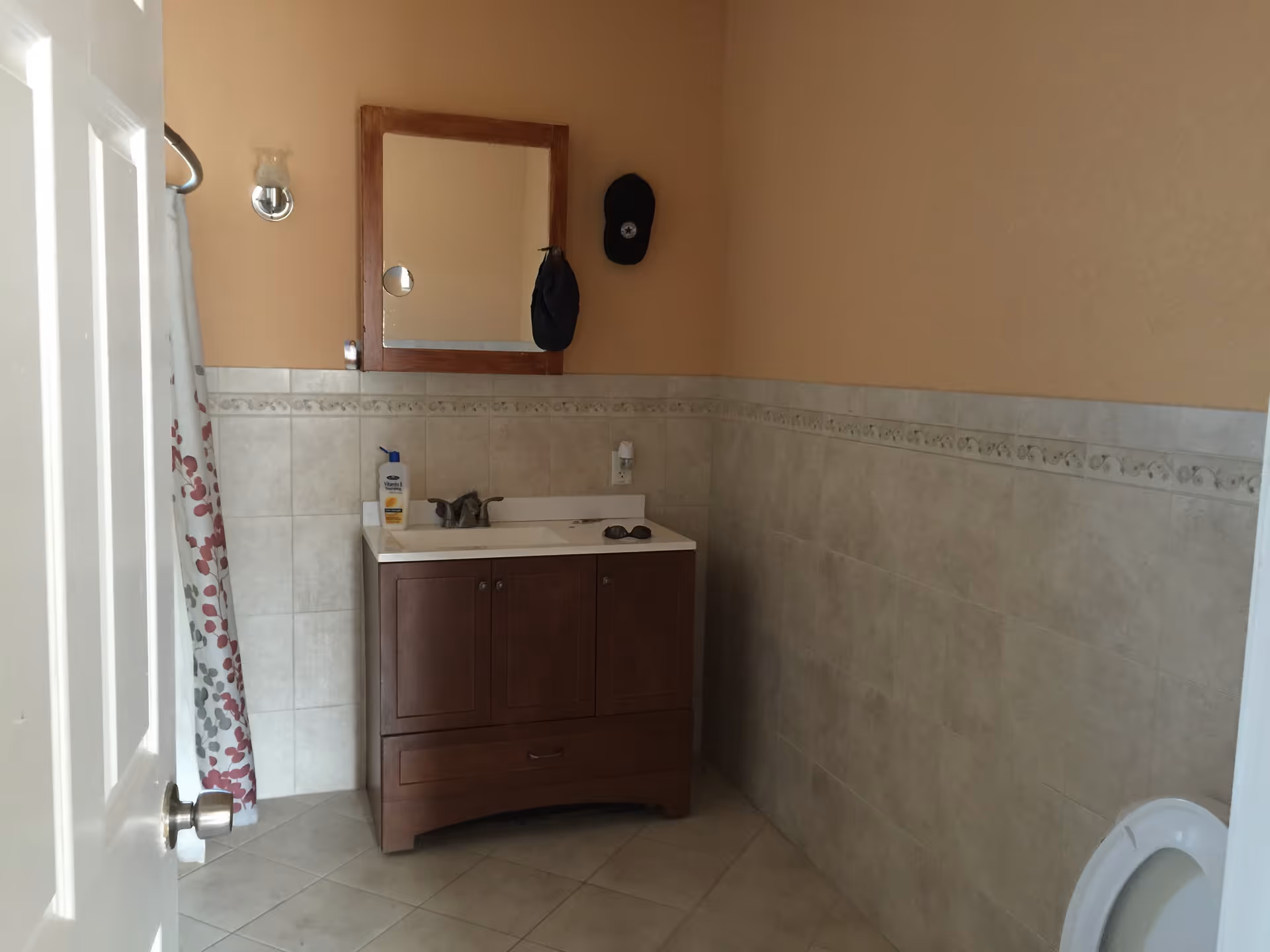 A bathroom with beige tiled walls and floor, a wooden vanity with a sink, a mirror above the sink, a shower curtain with a red and gray leaf pattern, and a partially visible toilet seat on the right side.