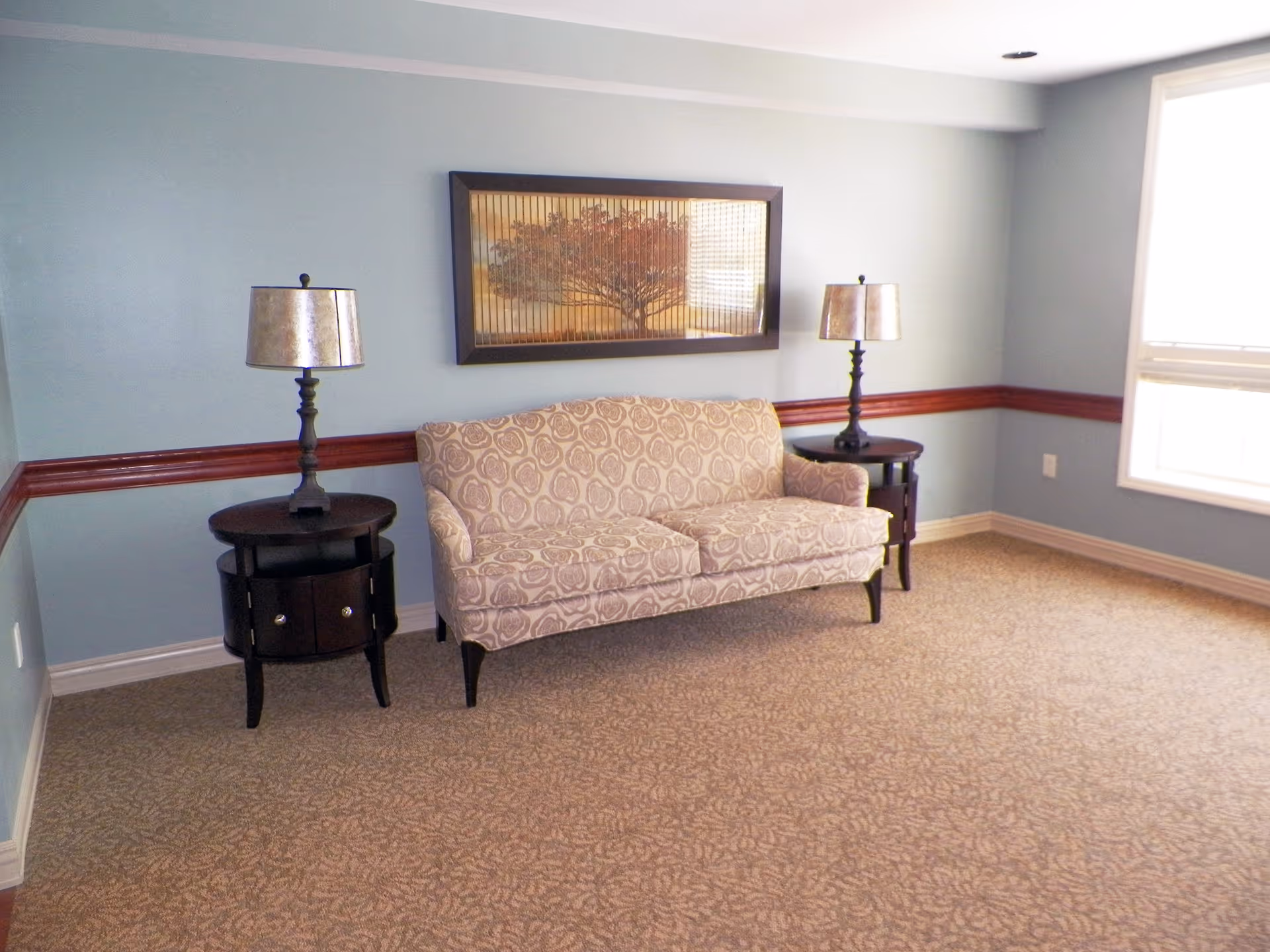A small sitting area with a patterned beige loveseat centered between two dark wooden side tables, each holding a lamp with a metallic shade. The walls are painted light blue with a dark wood chair rail, and a framed artwork depicting a tree hangs above the loveseat. A large window on the right side lets in natural light, illuminating the carpeted floor.