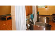 Interior view of a healthcare facility room with two stainless steel bathing tubs separated by a curtain. The room has tiled floors and walls, a wooden cabinet above the tubs, and a bench in the background.