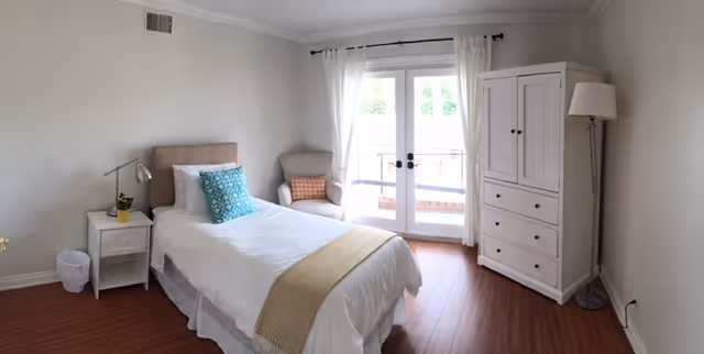 A bright and clean bedroom with a single bed made with white linens, a beige throw blanket, and decorative pillows. Next to the bed is a white nightstand with a small plant and a lamp. There is a comfortable armchair with a cushion near glass double doors with white curtains that lead to an outdoor area. A white wardrobe and a tall floor lamp are positioned against the right wall. The room has wooden flooring and light-colored walls.