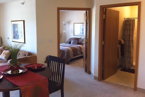 View of an interior living space showing a dining table with chairs and a red table runner in the foreground, a living room area with a couch and framed artwork on the wall to the left, a bedroom with a bed and lamp visible through an open doorway in the center, and a bathroom with a shower curtain and towels visible through an open door on the right.