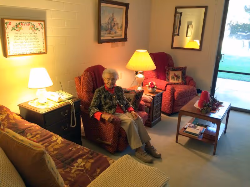 An elderly woman sitting in a red armchair in a cozy living room with warm lighting. The room features another matching armchair, a wooden coffee table with decorative items, a side table with a lamp and telephone, a couch with patterned cushions, framed artwork on the walls, and a large window or glass door showing a green outdoor area.