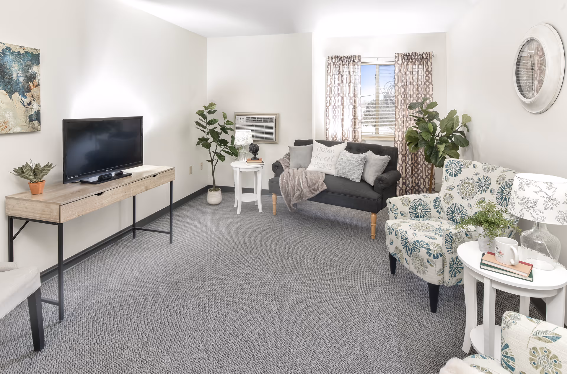 A bright and cozy living room with a gray carpet, a black loveseat with decorative pillows and a throw blanket, a patterned armchair, two white side tables with lamps and plants, a flat-screen TV on a wooden console table, a large wall clock, and a window with patterned curtains letting in natural light.