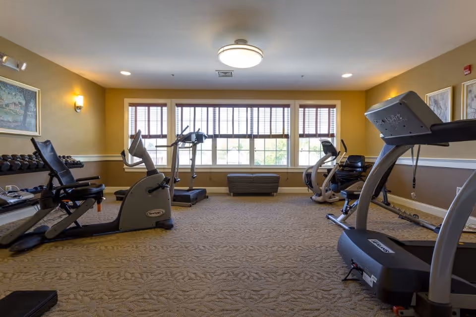 A fitness room with exercise equipment including treadmills, stationary bikes, and a set of dumbbells. The room has large windows with blinds allowing natural light to enter, beige walls with white trim, carpeted floor, and framed artwork on the walls.