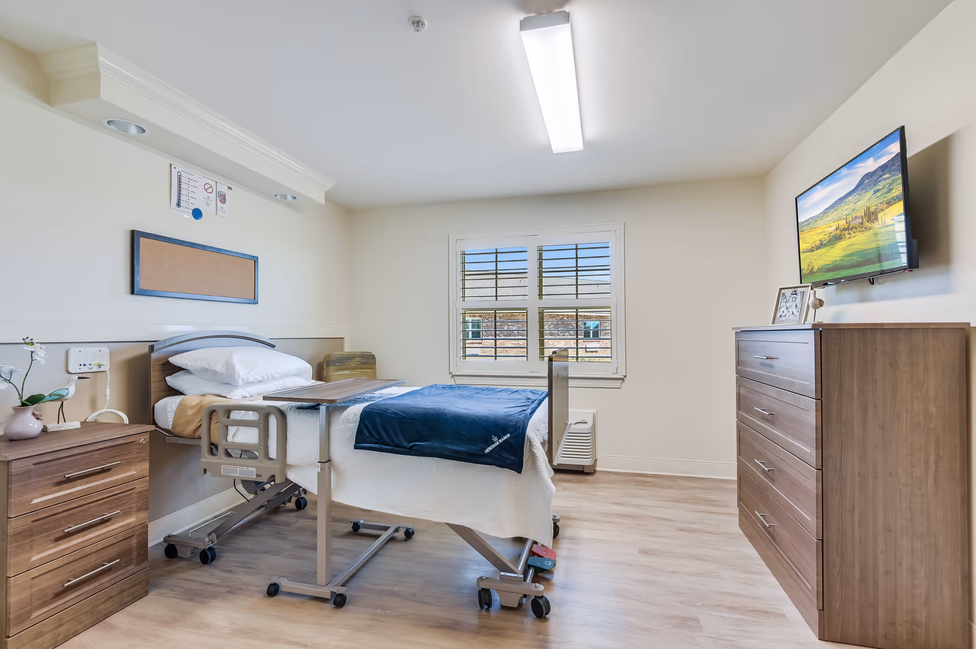 A tidy senior care resident bedroom with a hospital-style bed, bedside table, dresser, wall-mounted TV, and a window with shutters.