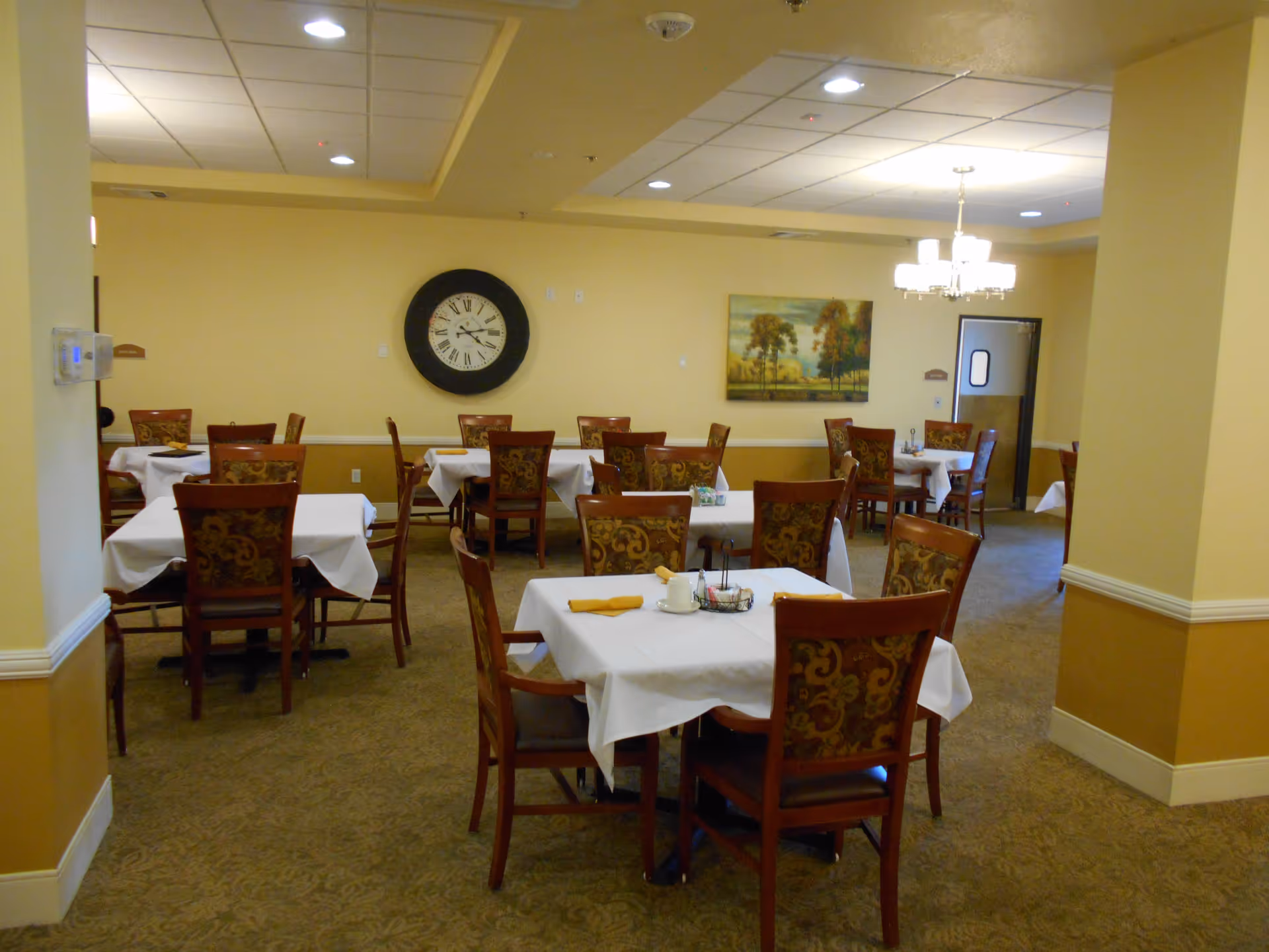 Dining room with multiple tables covered with white tablecloths, each surrounded by wooden chairs with patterned upholstery. A large round clock and a landscape painting hang on the beige walls. A chandelier is visible on the ceiling, and the room has a carpeted floor.