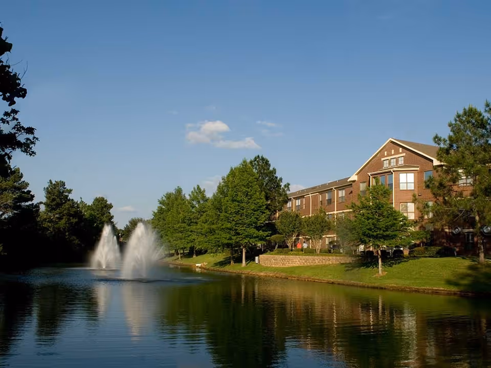 A serene outdoor scene featuring a pond with two water fountains, surrounded by green trees and grass. To the right, there is a large brick building with multiple windows under a clear blue sky.